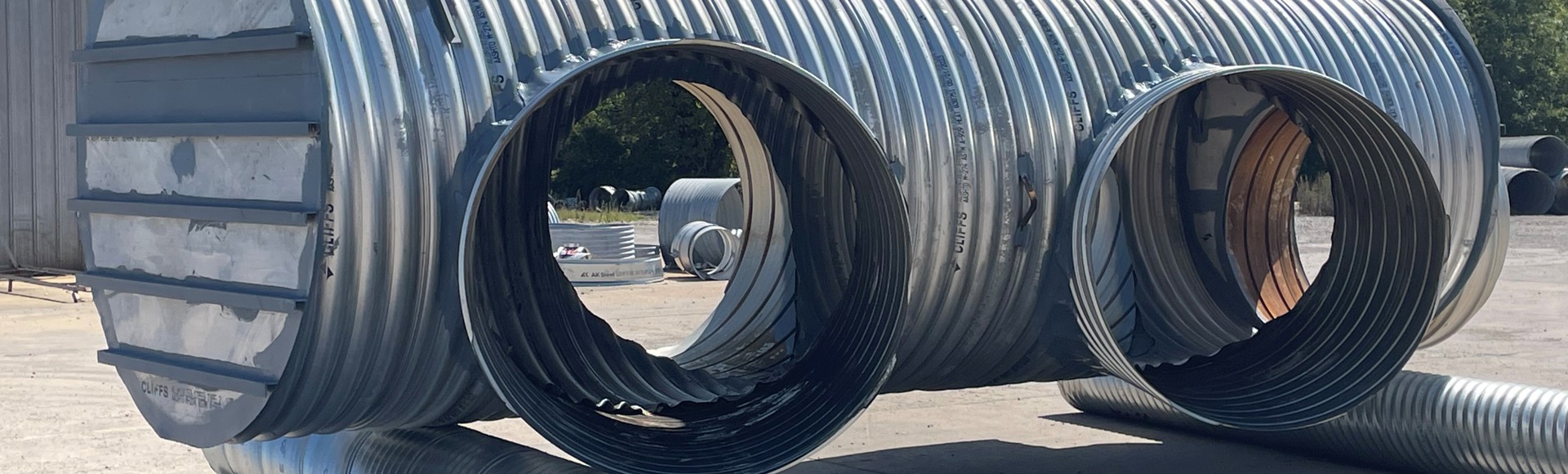 Large corrugated metal pipes assembled into a structure on a sunny day, with trees and building in the background.
