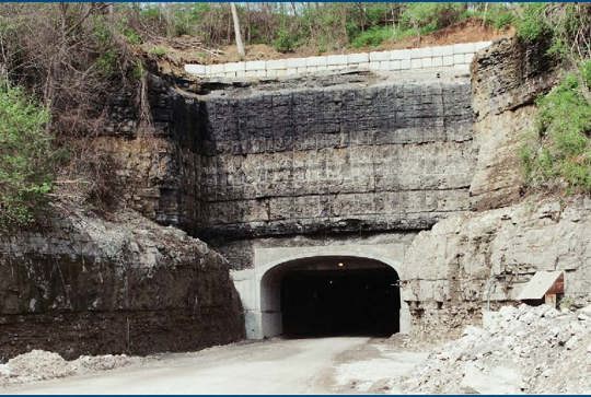 A wide tunnel entrance set into a rocky, layered hillside, surrounded by trees and vegetation.