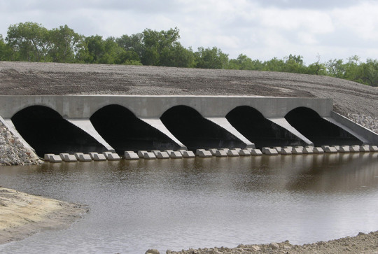 A water drainage structure with five arched concrete culverts spans a body of water, with earthen embankments on either side.
